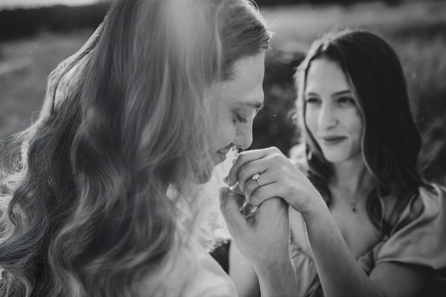 black and white portrait of a groom kissing his fiance's hand and her diamond engagement ring as she smiles at him