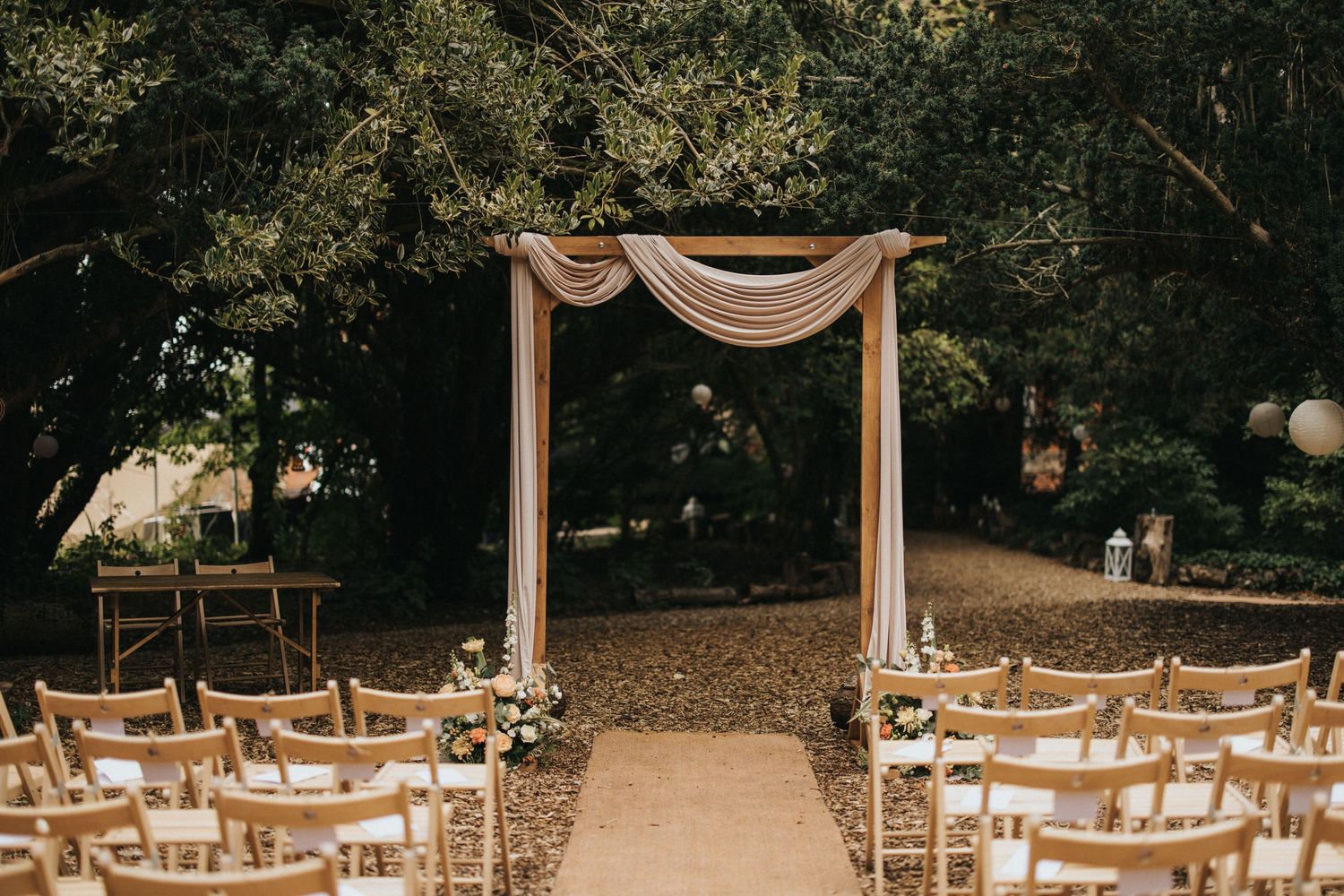A wooden wedding arch with white fabric draping stands at the end of an aisle lined with white chairs in a wooded setting at Hirst Priory.
