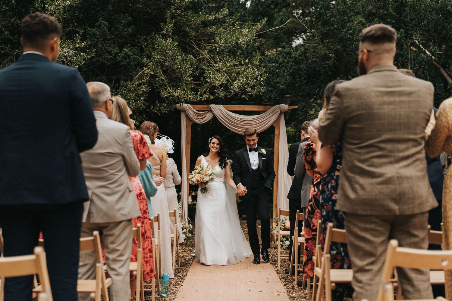 Guests stand and turn to watch the bride walk down an outdoor aisle at a garden wedding ceremony at Hirst Priory.