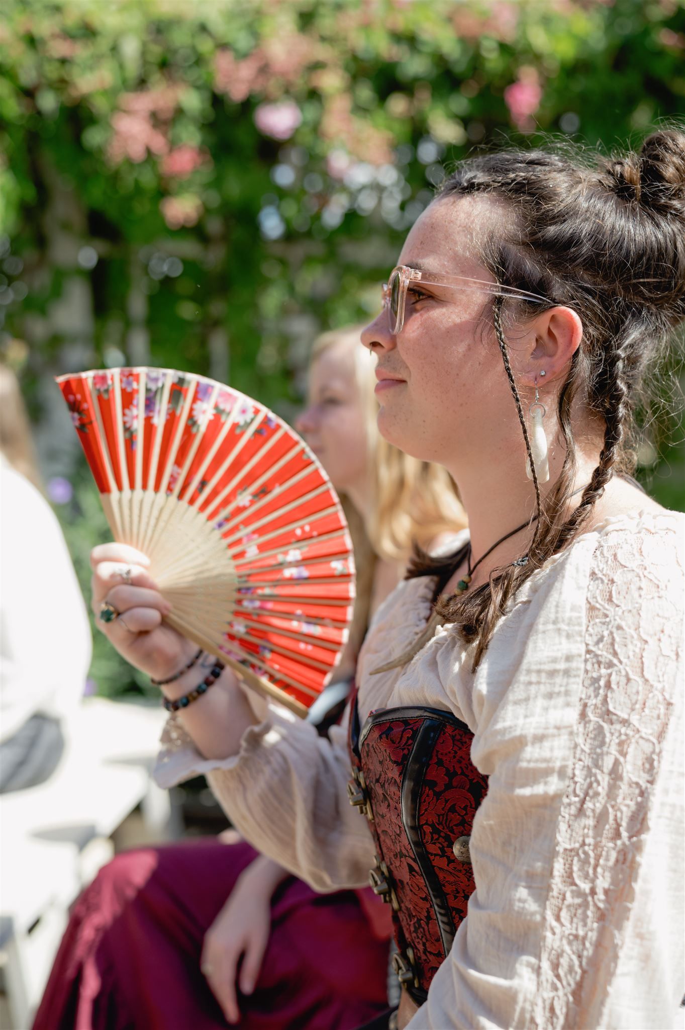 Performers in vintage costumes hold decorative red and white handheld fans while gathered outdoors under flowering trees.