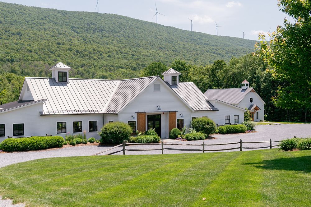 White barn-style buildings with wood fencing set against green mountain backdrop and wind turbines on the ridge line.