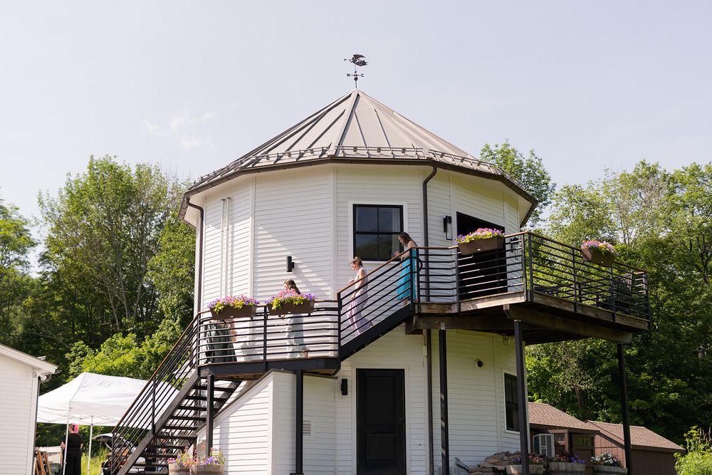 Two people stand on the deck of a unique round lighthouse-style building with metal stairs against a lush green backdrop.
