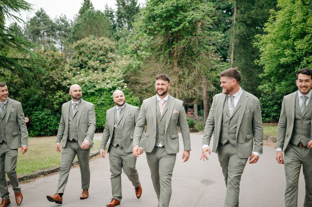 Groomsmen in matching light gray suits walk together on a path surrounded by lush green trees and gardens.
