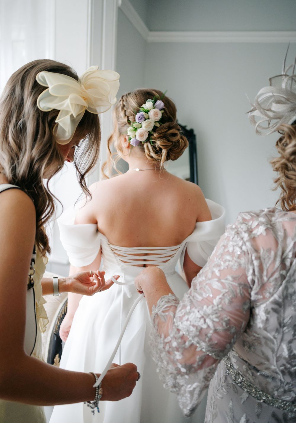 Wedding party helps with dress preparations showing delicate lace details and floral accessories in soft grey tones.