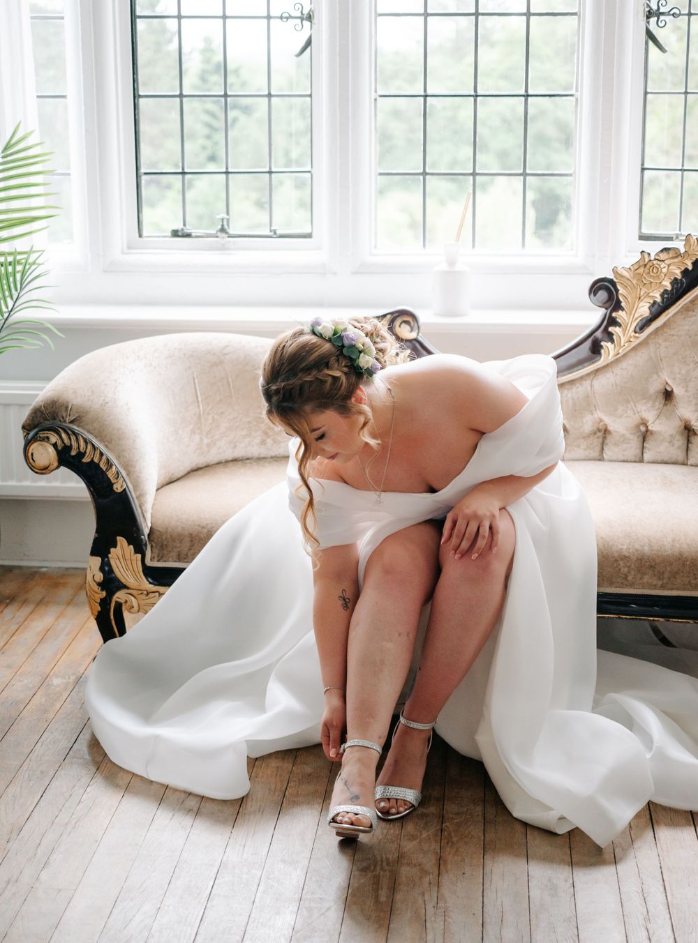 Bride in white gown sits on vintage couch adjusting her strappy wedding shoes near bright windows.