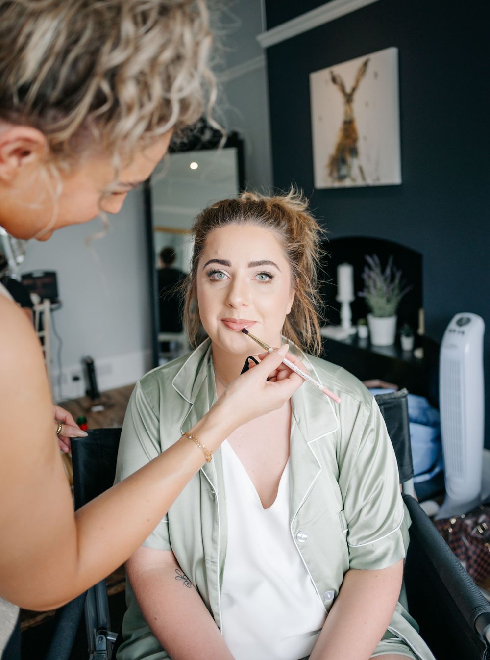 A makeup artist applies cosmetics to a client wearing a sage green shirt in a room with dark walls and artwork.