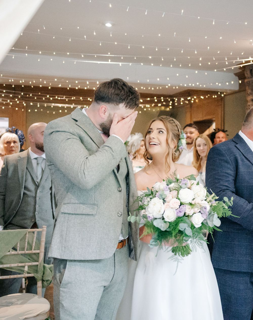 An emotional moment between a couple at their wedding ceremony under twinkling lights while holding a white floral bouquet.