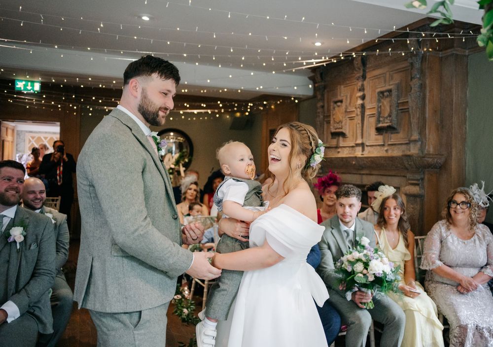 A couple sharing a tender moment with a baby during their rustic barn wedding ceremony with string lights overhead.