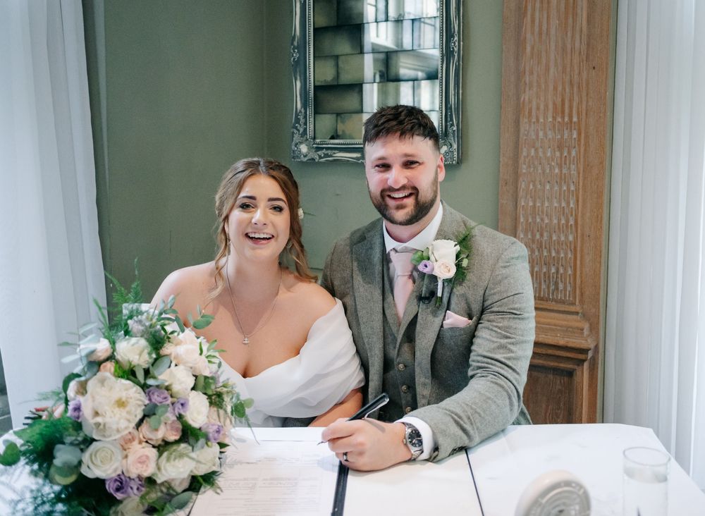 A couple sits together at a table during their wedding, with a soft pink and white floral arrangement in the foreground.