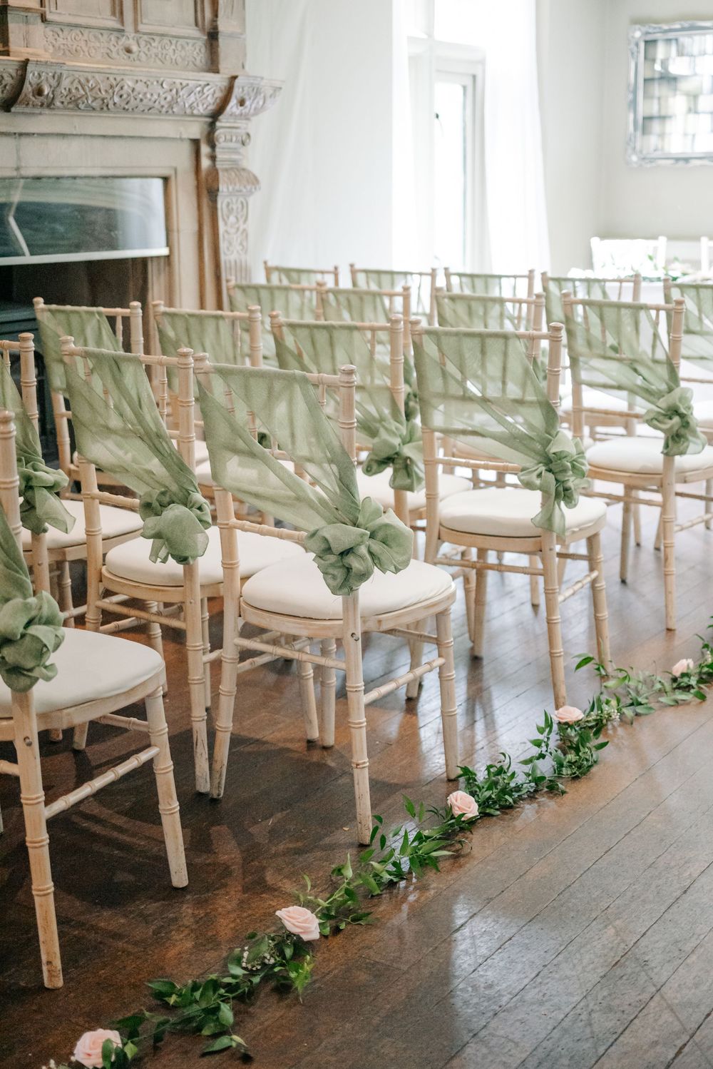 Elegant wedding ceremony chairs with sage green sashes and greenery aisle decorations in a bright indoor venue.