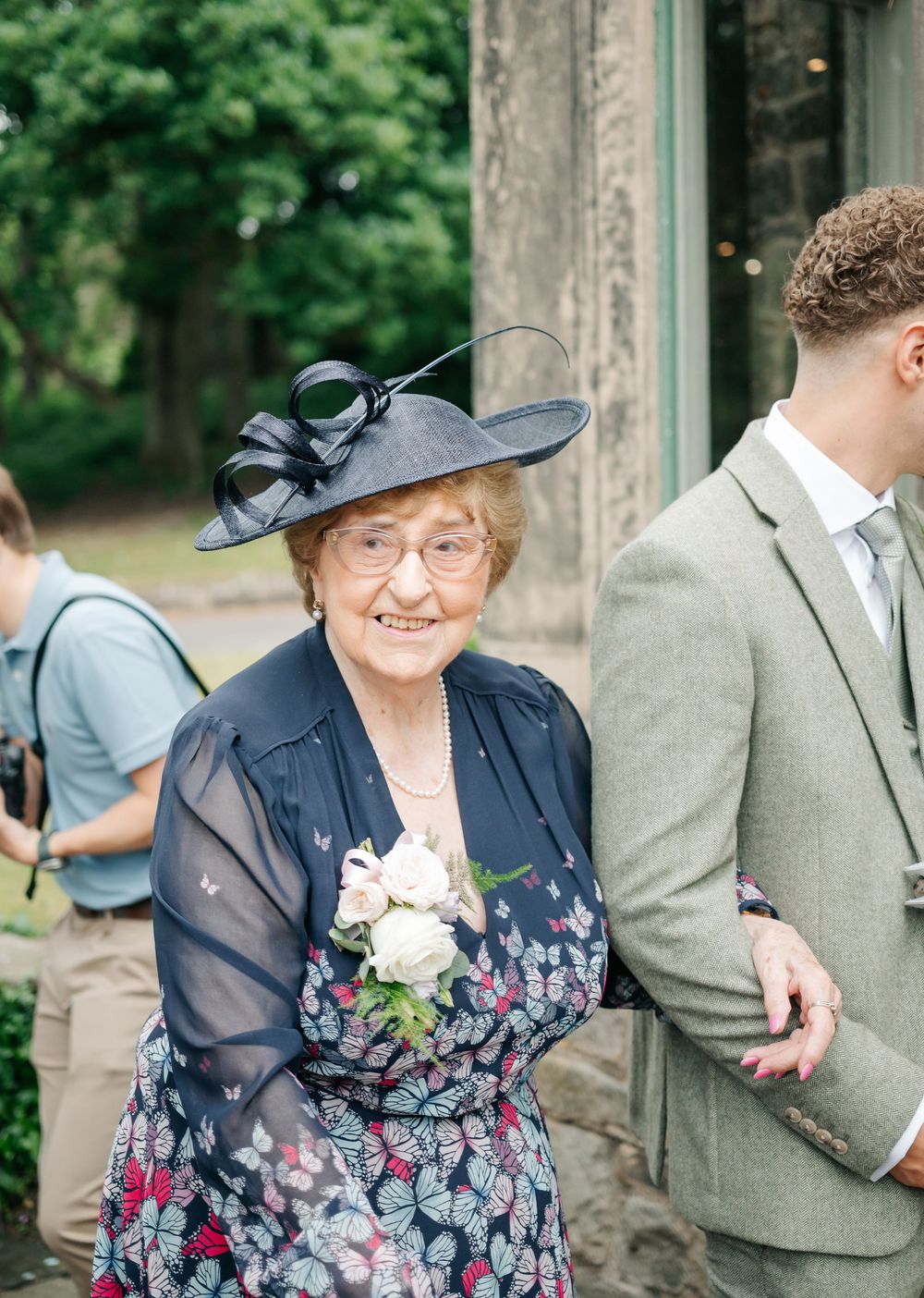 Wedding guest wearing a navy dress and formal black hat with bow detail standing outdoors.