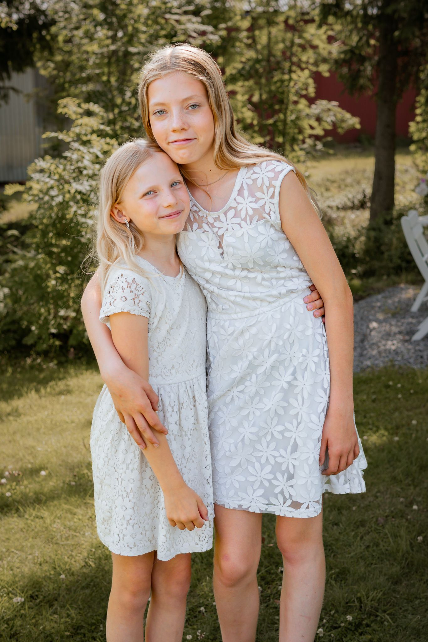 Två tjejer i vita spetsklänningar håller om varandra i en solig trädgård en sommardag. Familjefotograf, Umeå, Fotograf, Västerbotten