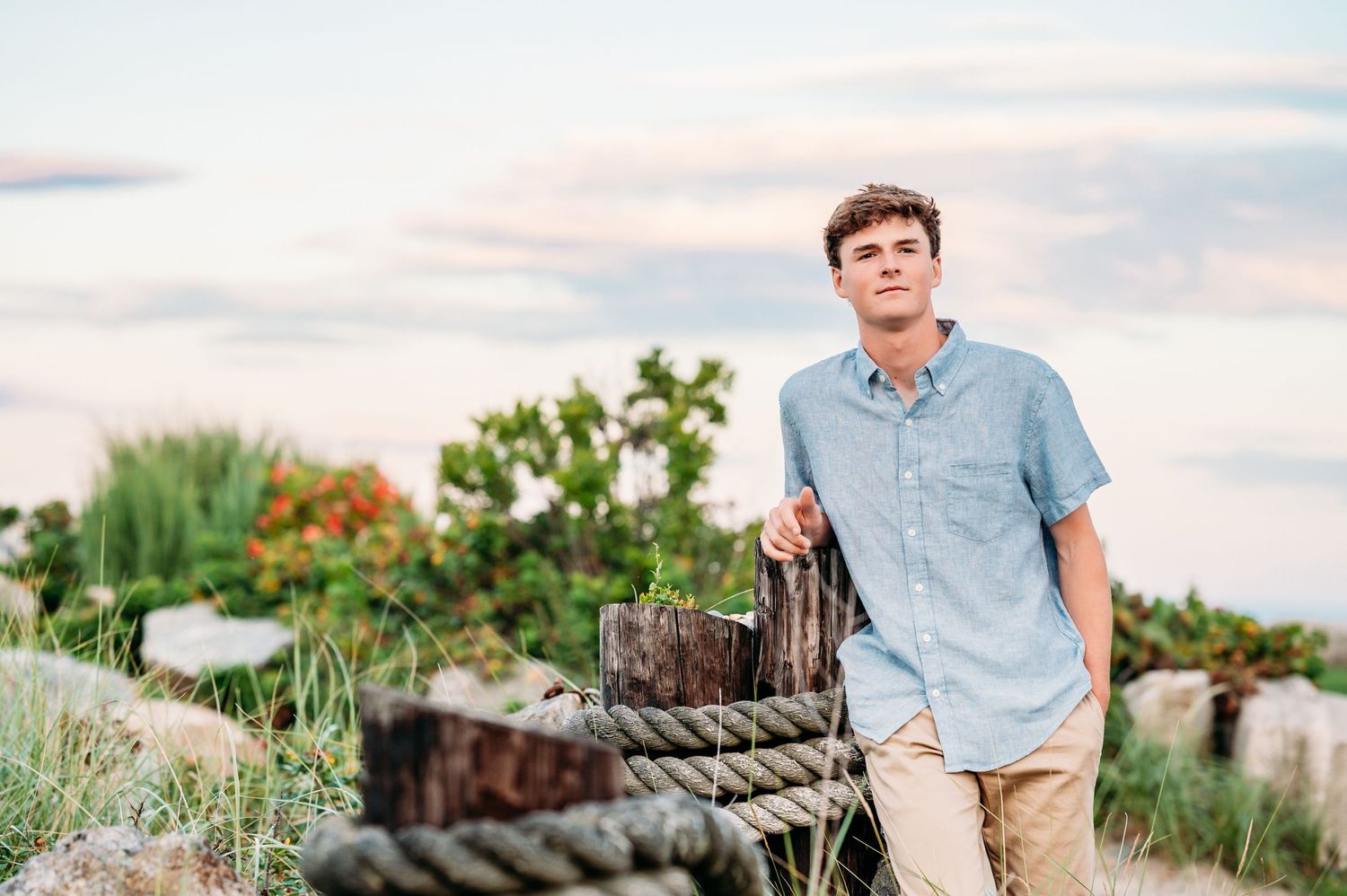 Someone in a light blue shirt and khaki pants poses outdoors near wooden beams and greenery at sunset.