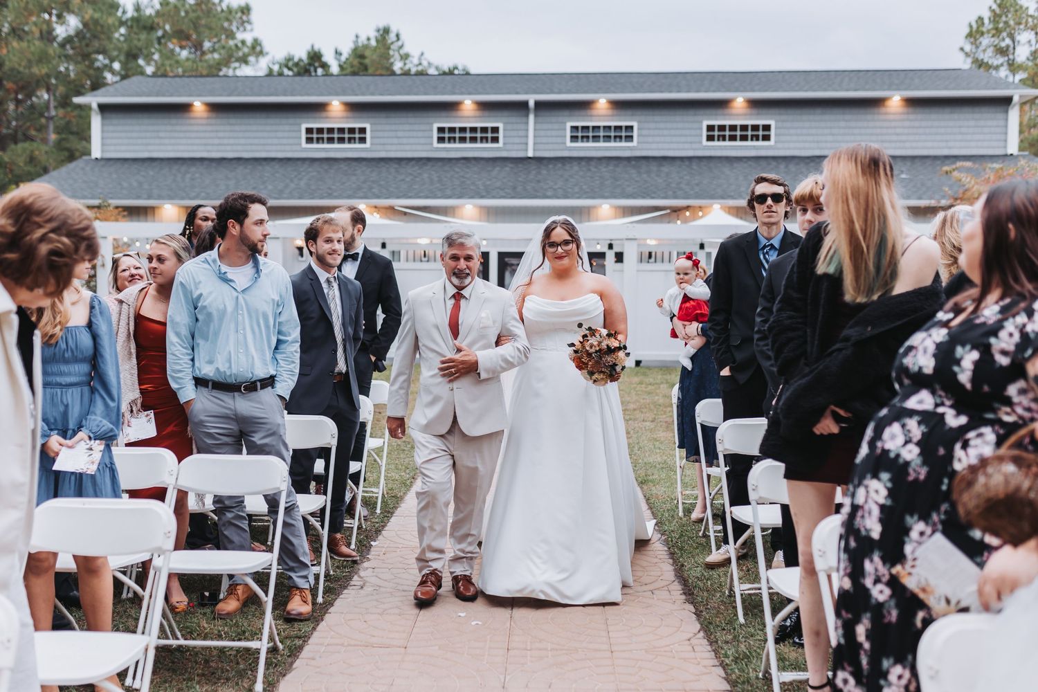 Outdoor wedding ceremony at a rustic barn venue with guests seated along a petal-lined aisle.