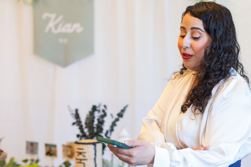 A person with long curly dark hair wearing a white shirt stands smiling while looking at their phone near a lavender plant.