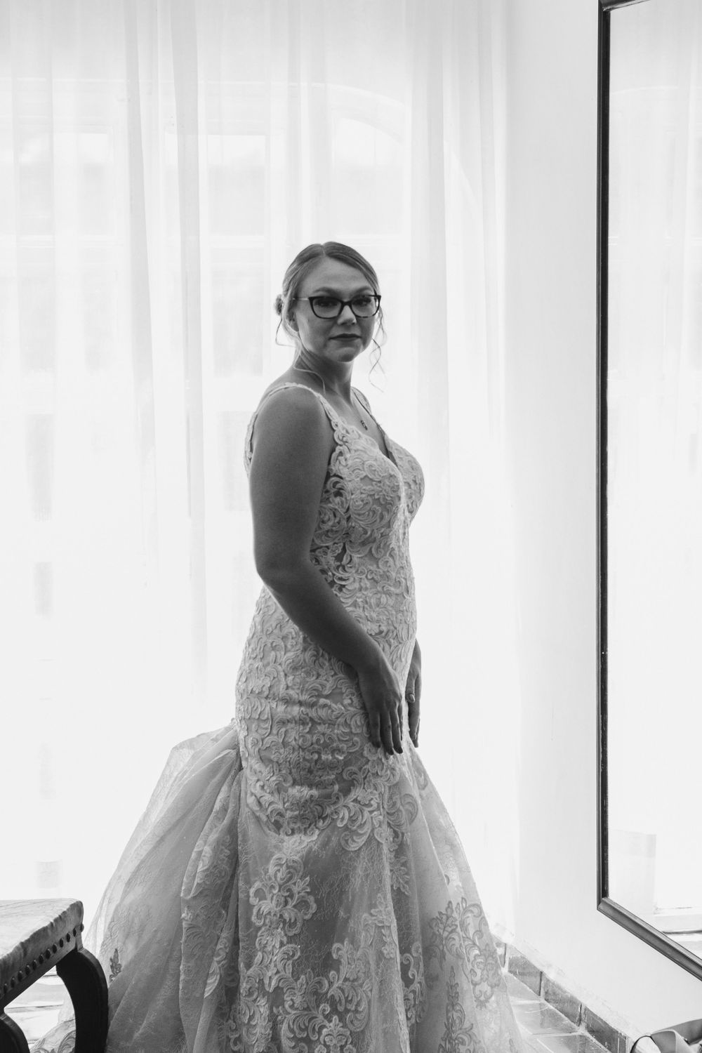 A person in a detailed lace wedding dress poses near a window in a black and white photograph.