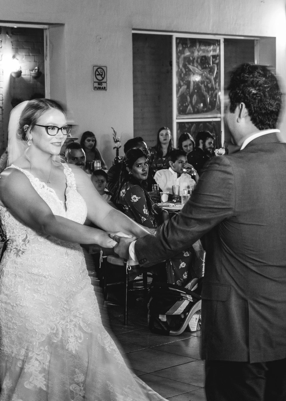 A black and white photograph showing a couple dancing together at a wedding reception with guests watching in the background.