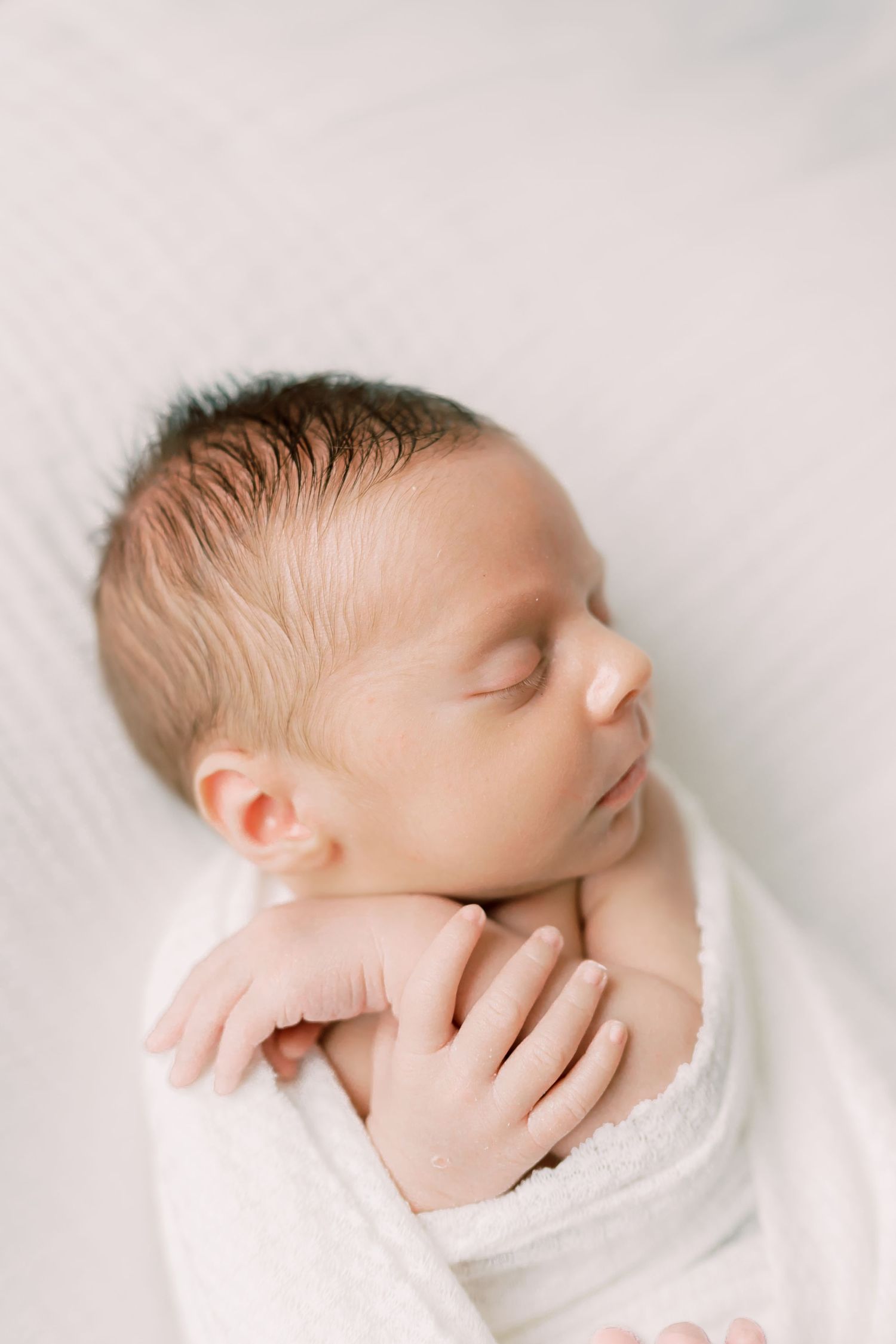 Sleeping newborn baby wrapped in white blanket against soft white background in minimalist portrait style.