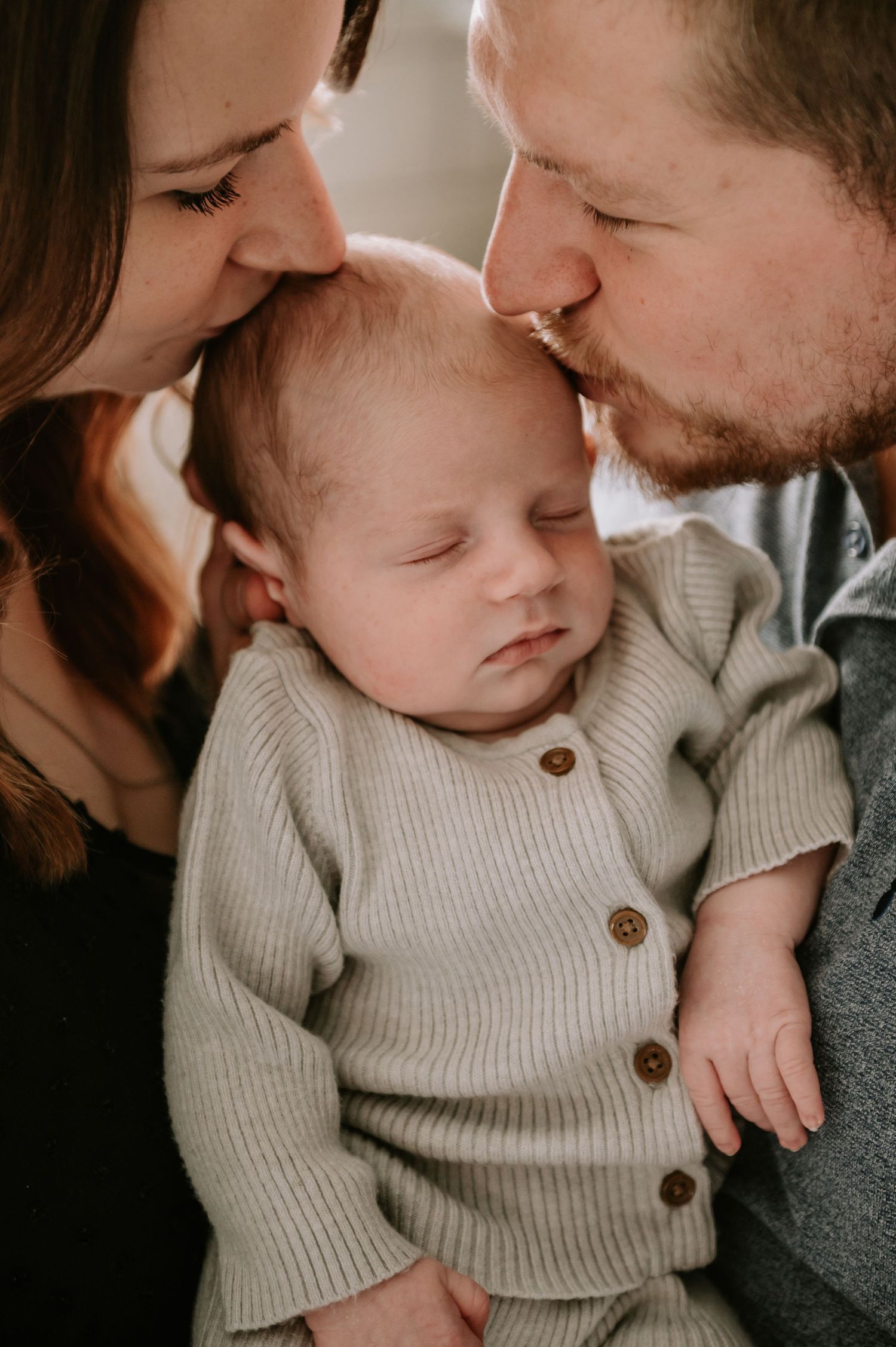 A sleeping baby wearing a beige cardigan is tenderly embraced by loving parents in an intimate family portrait.