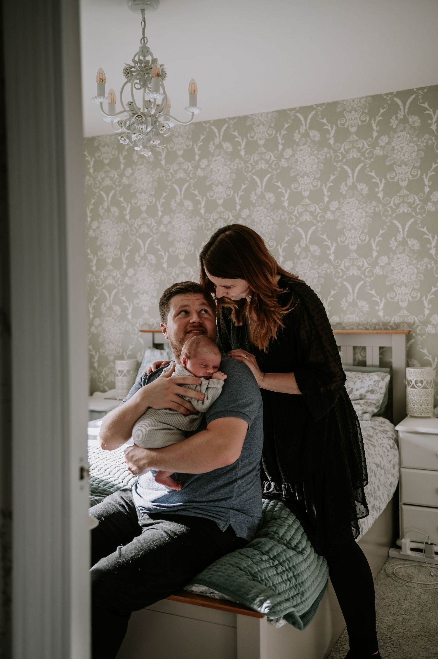 Cozy nursery scene with floral wallpaper and elegant chandelier lighting creating an intimate family moment.