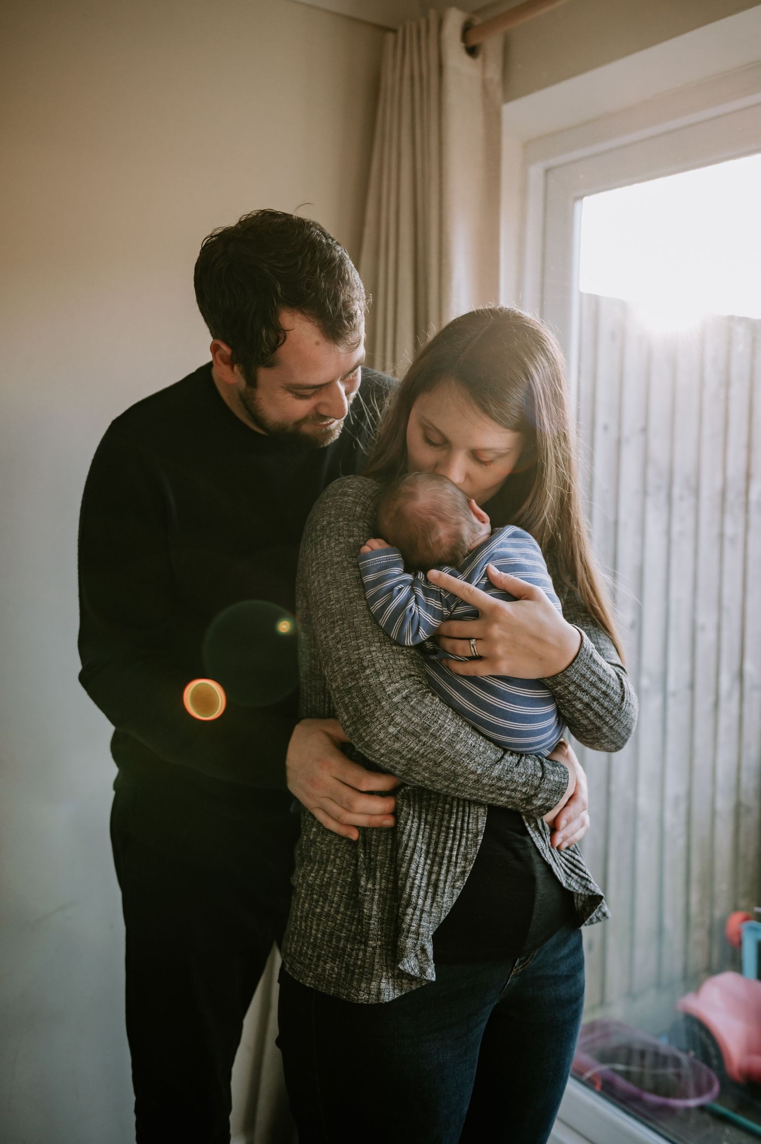 A couple stands by a window holding their baby in a cozy indoor setting with natural light streaming in.