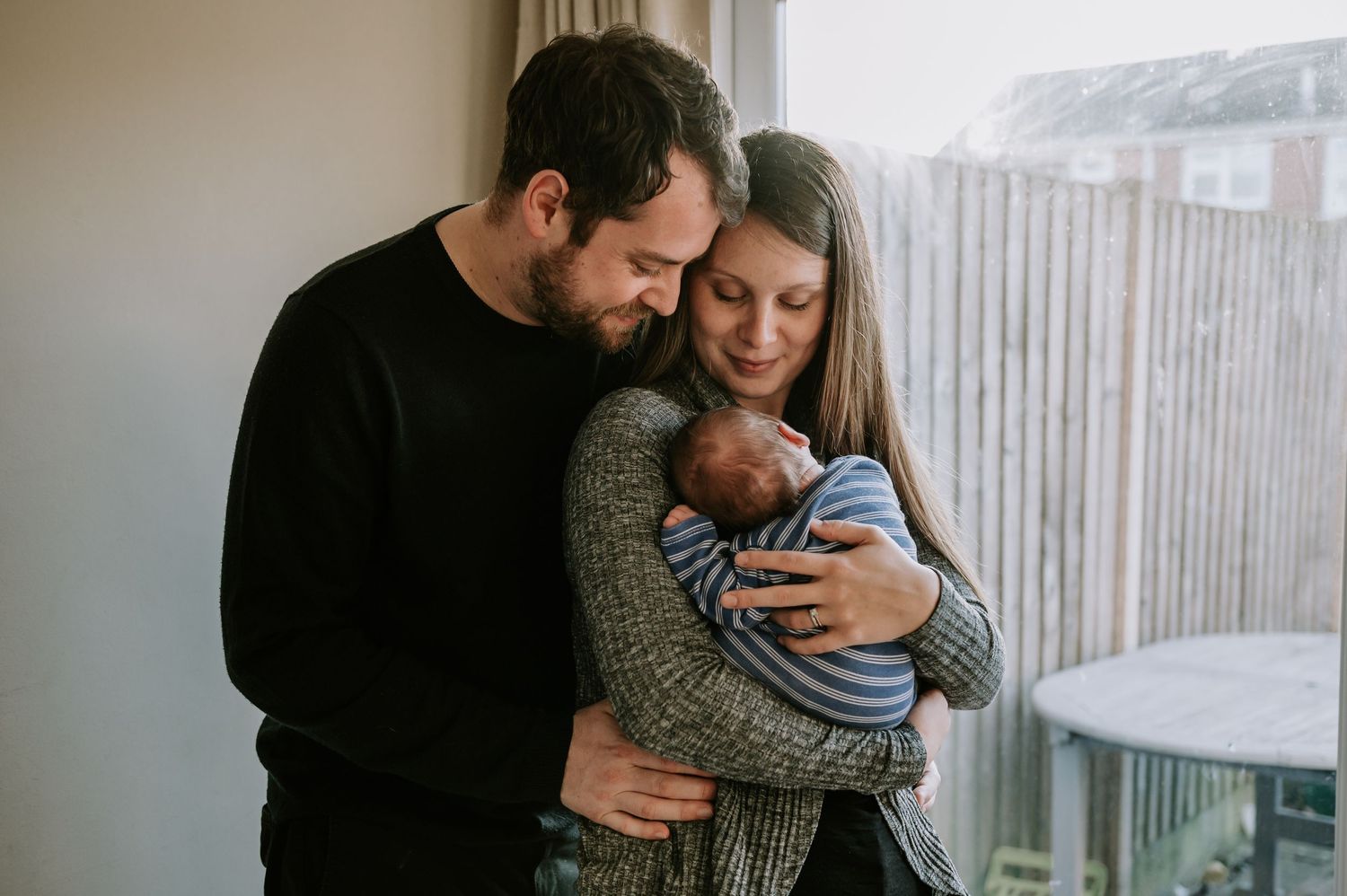 A couple lovingly embraces their newborn baby near a window in a tender family moment.