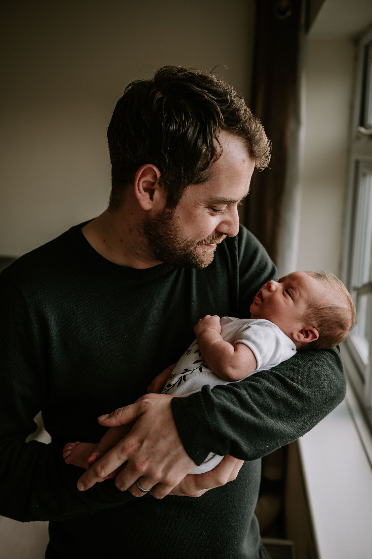 A tender moment near a window shows a person in a dark sweater gently cradling a newborn baby in their arms.