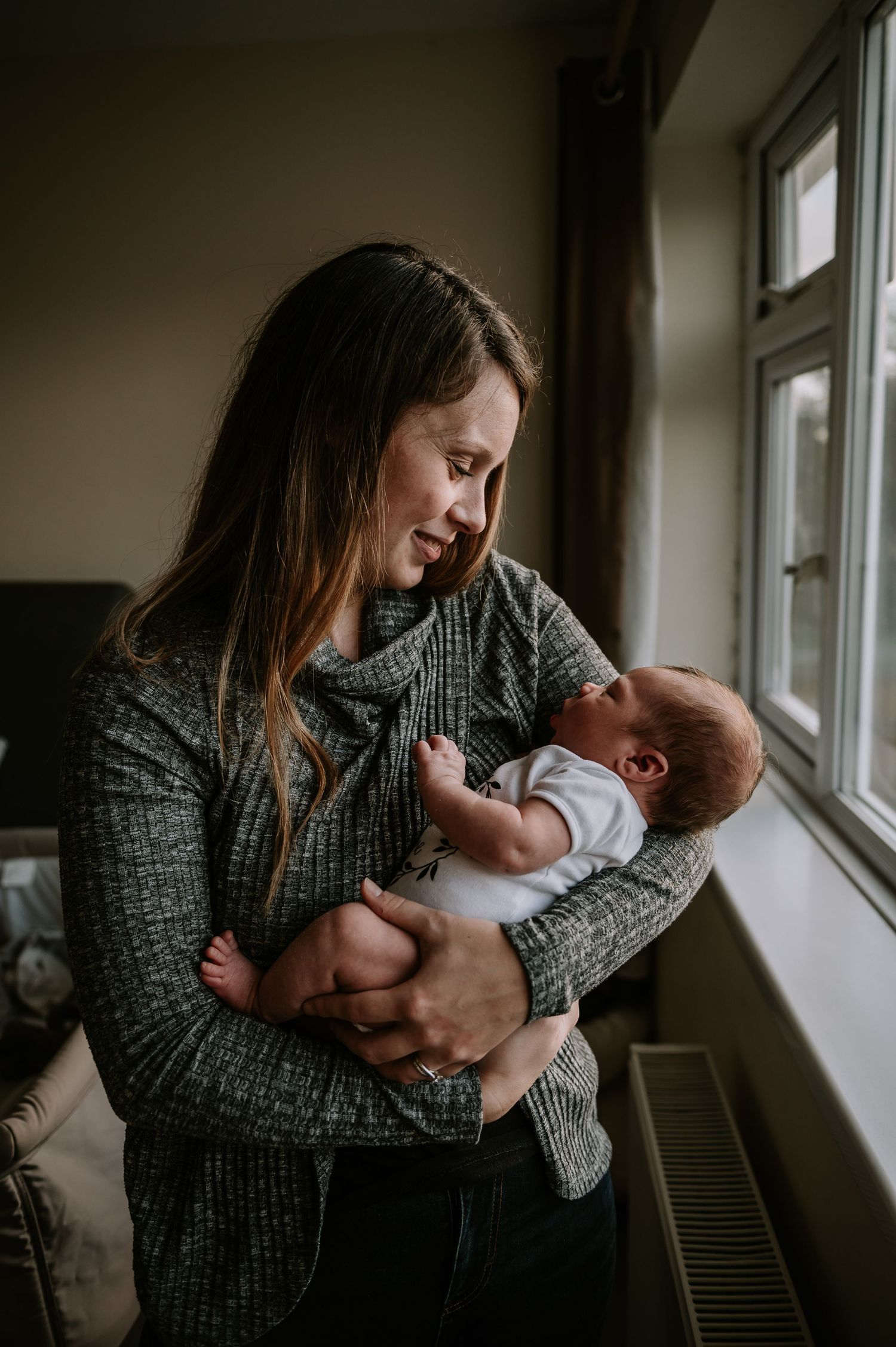 A parent in a gray sweater holds and gazes lovingly at a newborn baby next to a sunny window.