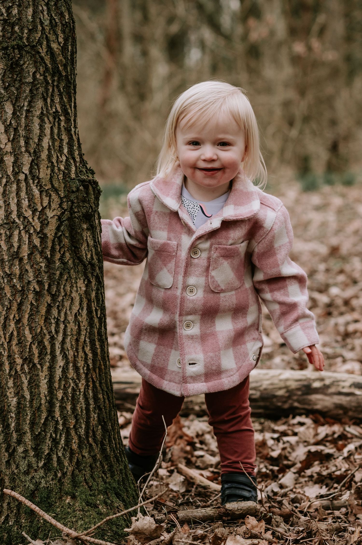 A small child wearing a pink plaid jacket and burgundy pants stands next to a tree trunk in autumn woods.