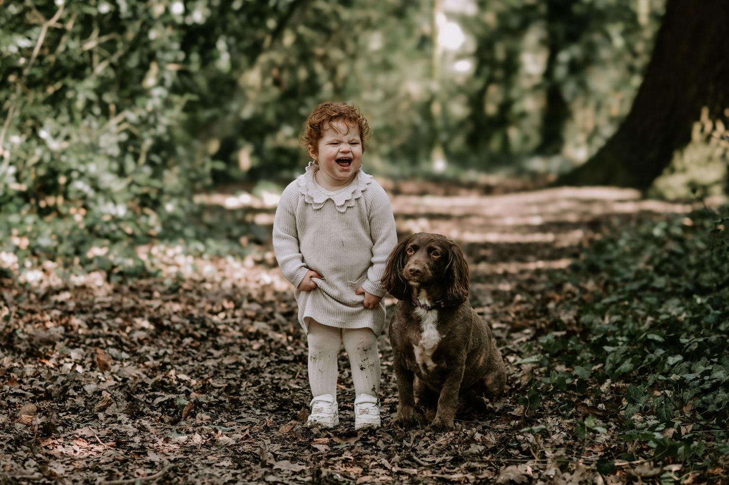 A small child in a beige outfit shares a joyful moment with their brown and white dog on a wooded path.