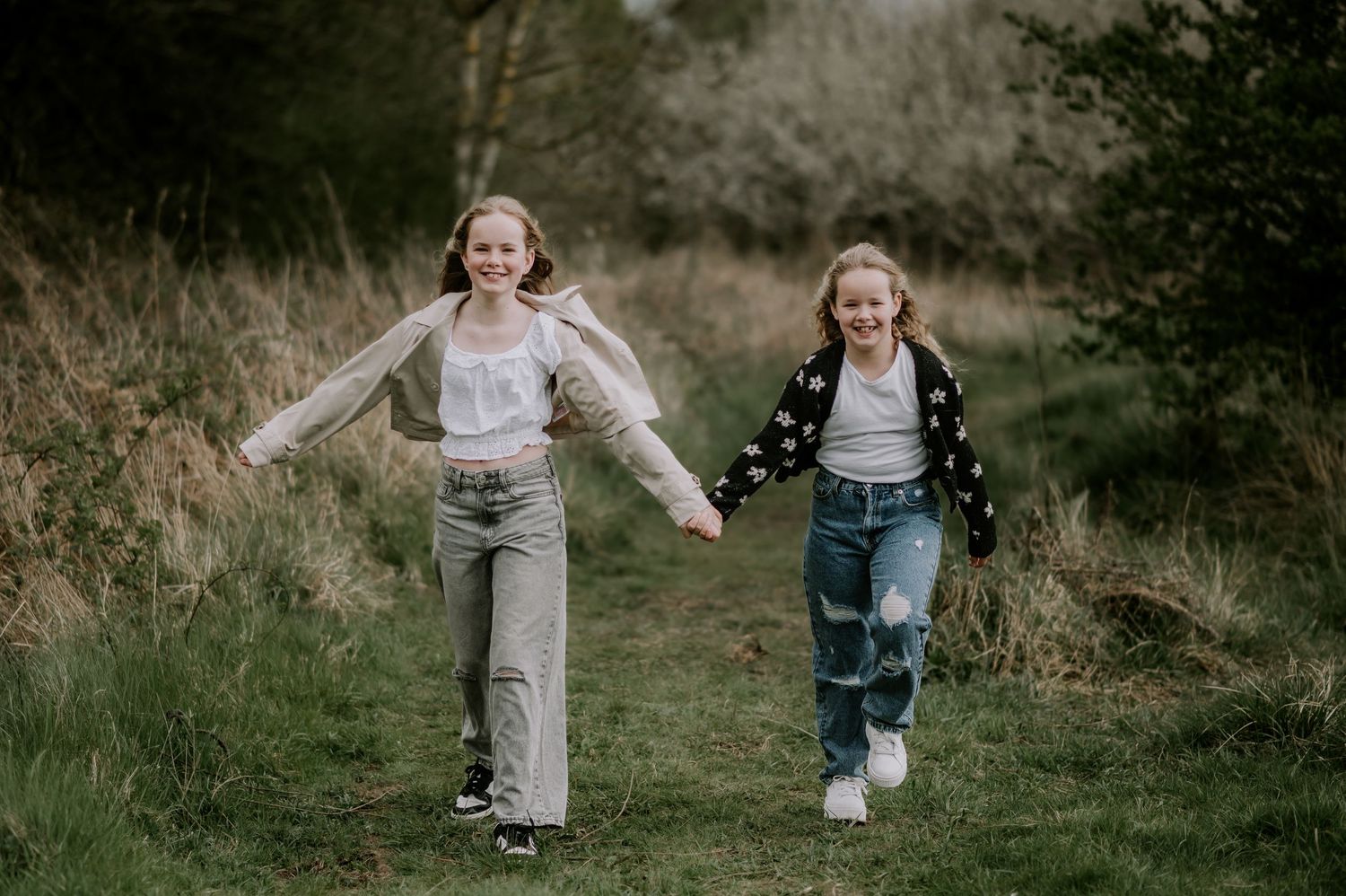 Two children in casual outfits holding hands and running joyfully together on a grassy path outdoors.