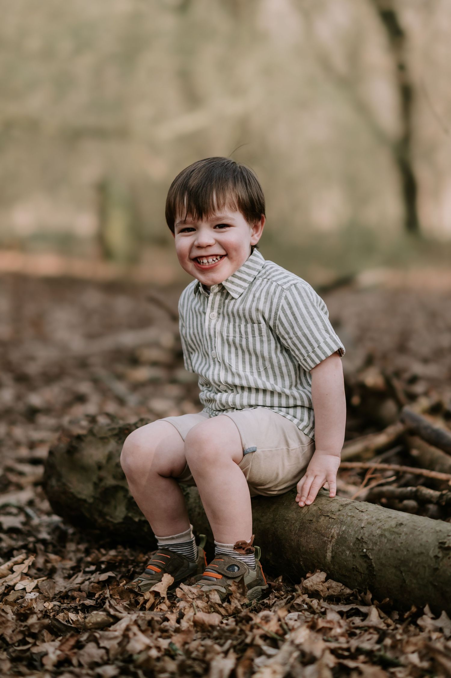 Small child in striped shirt and khaki shorts sits smiling on a fallen log surrounded by autumn leaves in a forest.