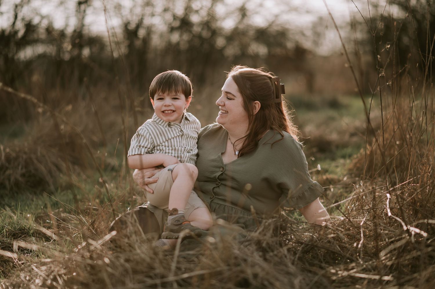 Two people share a joyful interaction while sitting in tall grass at sunset.