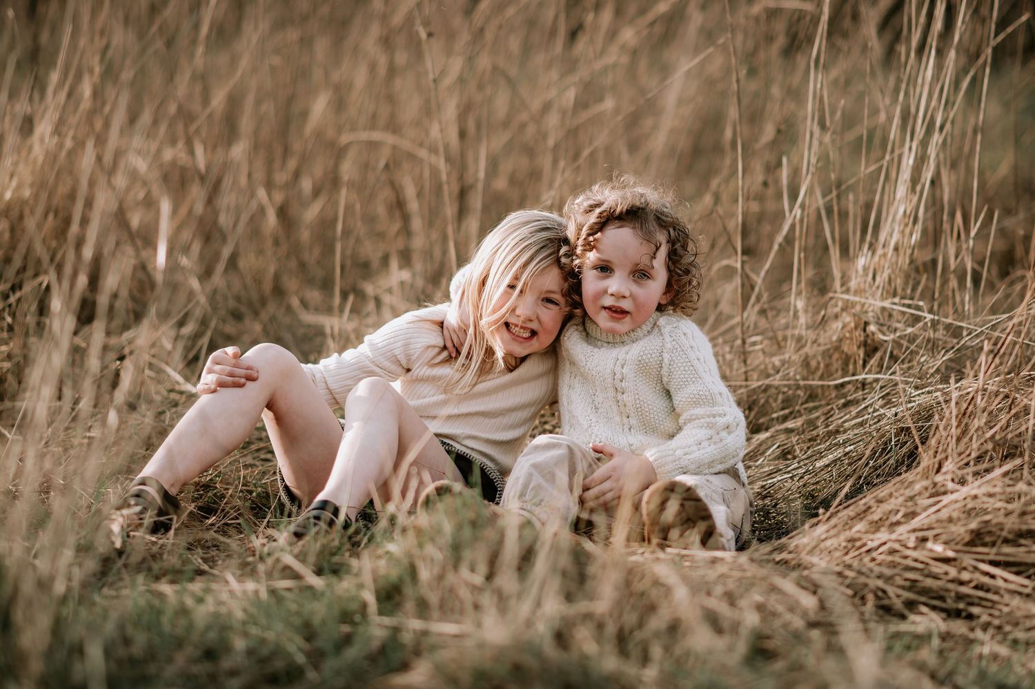 Two children sit together in golden tall grass on a sunny autumn day while laughing and playing.