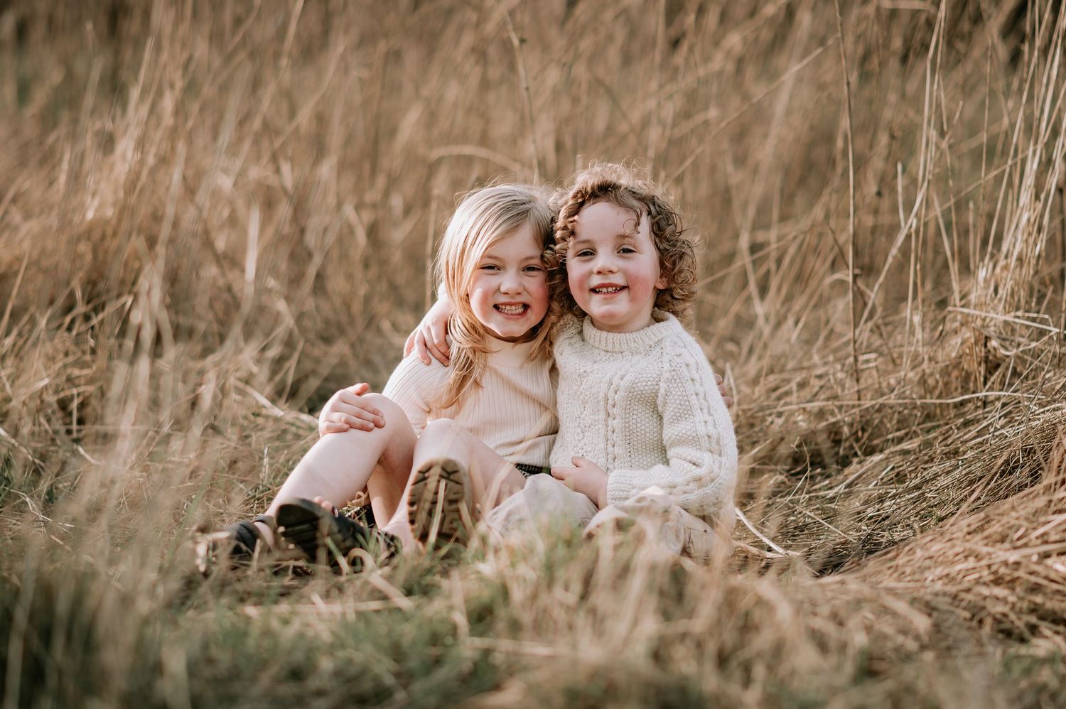 Two kids laughing together while sitting in tall golden grass during sunset.