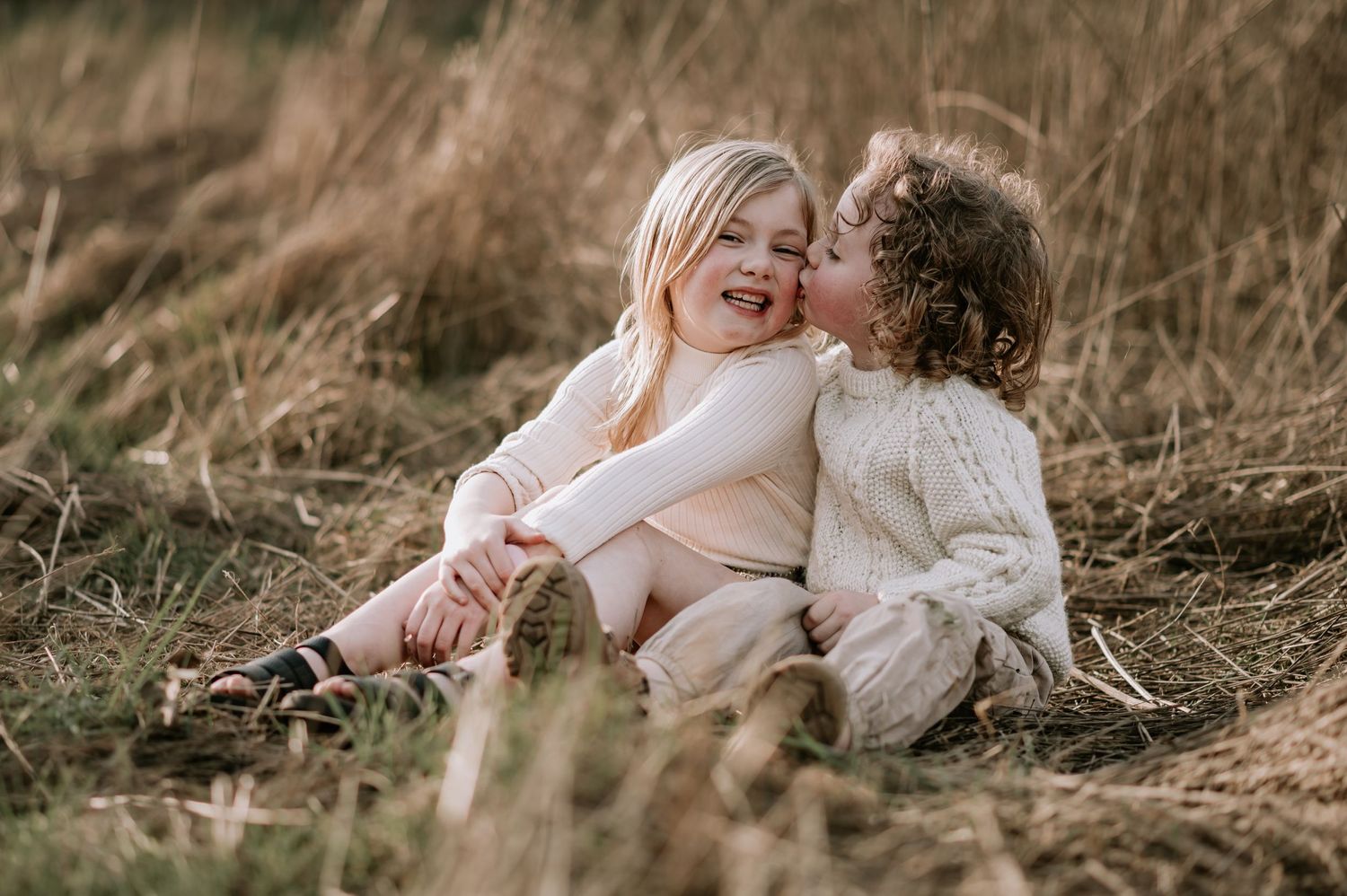 Two children in cozy sweaters sharing a sweet moment while sitting together in tall grass at sunset.