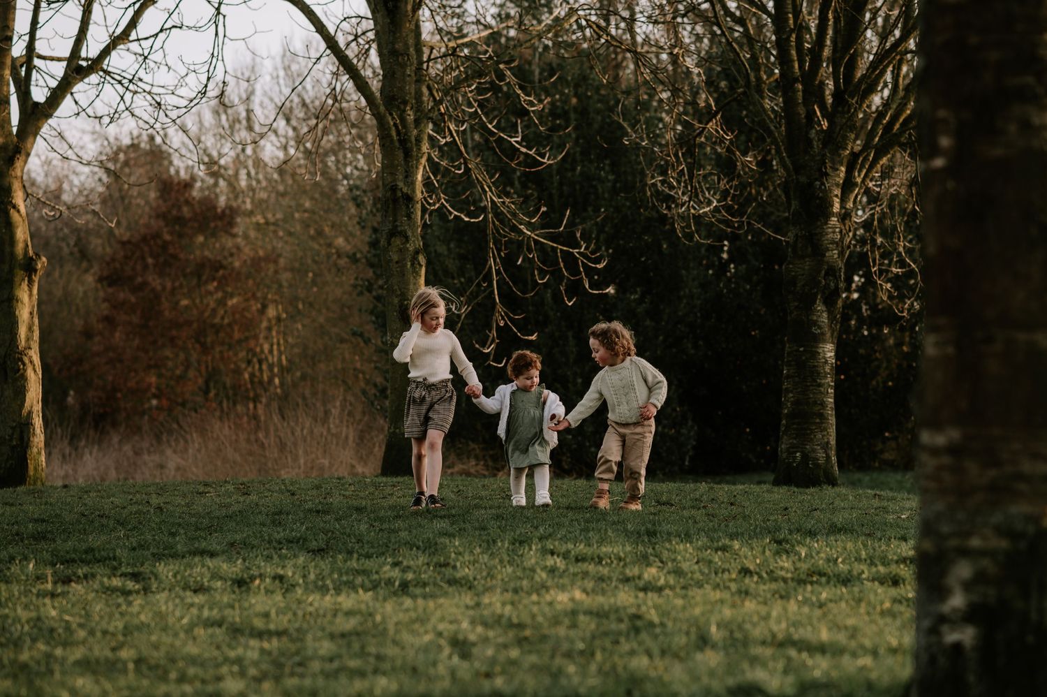 Three silhouettes running and playing together in a grassy field at sunset with trees in background.