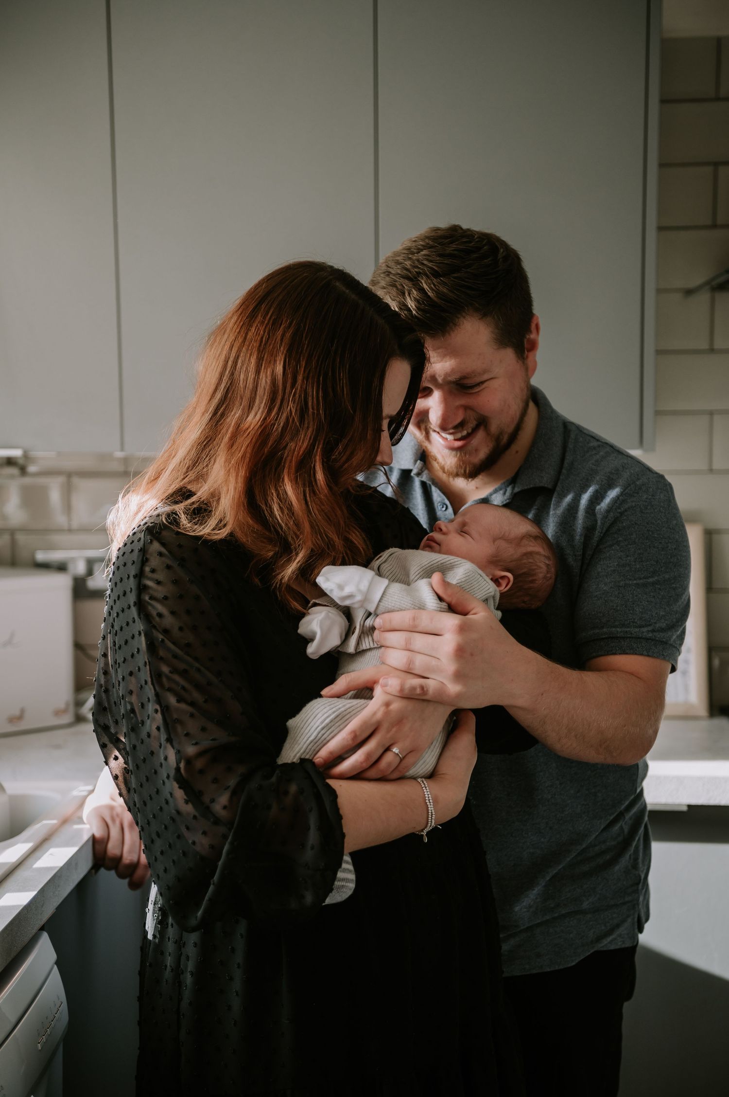 Two parents share an emotional moment while holding their newborn baby in a kitchen.