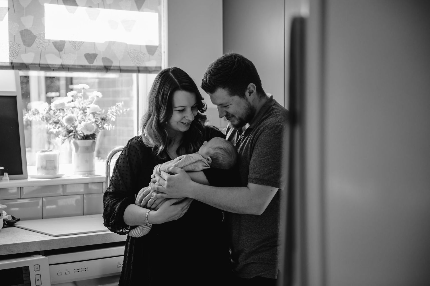 A tender intimate black and white moment captured in a kitchen as new parents share a moment with their newborn baby.