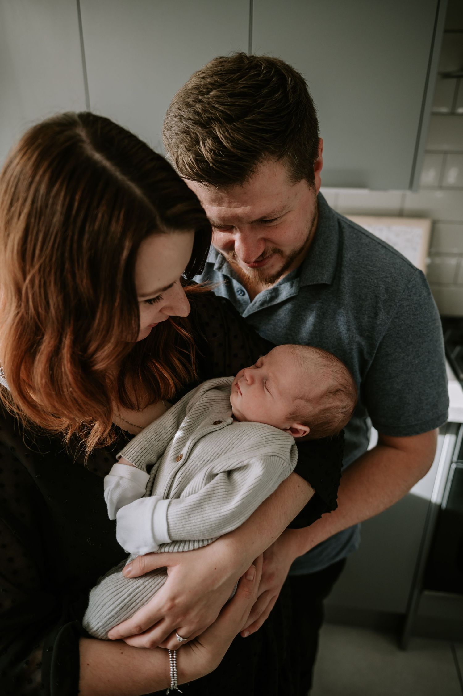 A couple gazes lovingly at their newborn baby wrapped in a gray blanket while standing in a room.