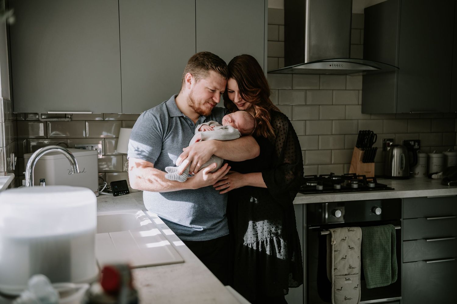 Parents hold their newborn baby in their modern kitchen while sharing an intimate moment together.