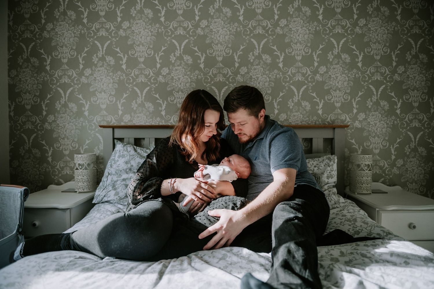 Parents lovingly embrace their newborn baby while sitting together on a bed with patterned wallpaper in background.