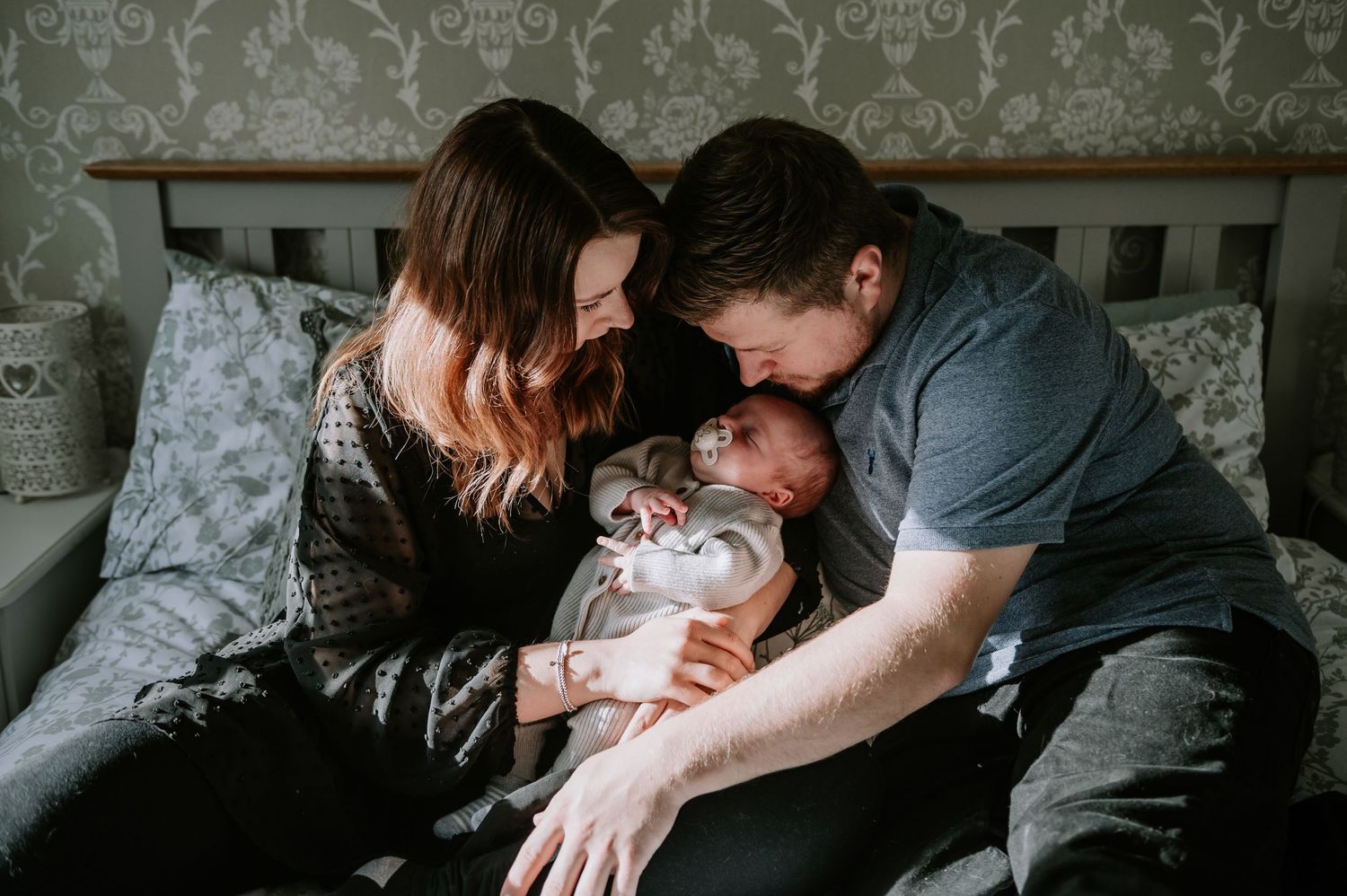 Parents lovingly embrace their newborn baby while sitting together on a couch with decorative pillows.