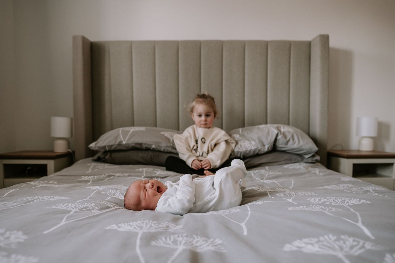 Small child sits cross-legged on a large bed with gray bedding and upholstered headboard.
