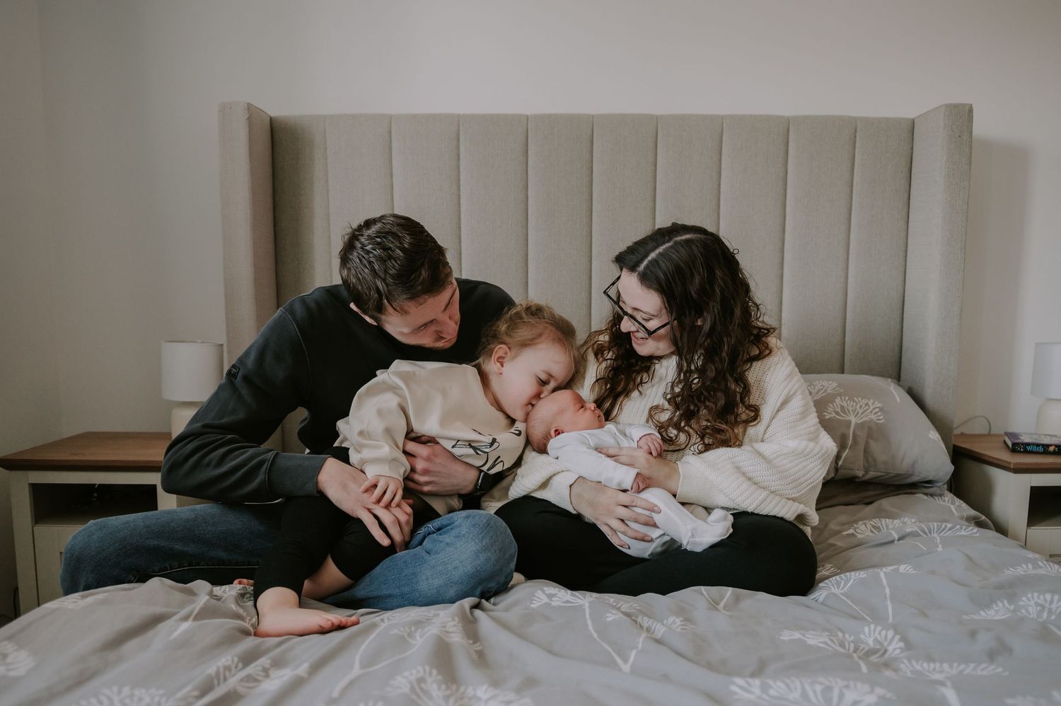Family snuggles together on gray bedding while bonding with their newborn baby.