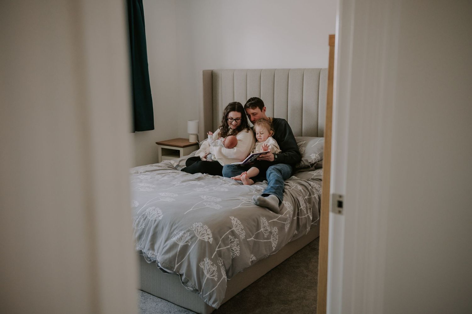 A couple sits together on a gray upholstered bed while looking at something on a digital device.