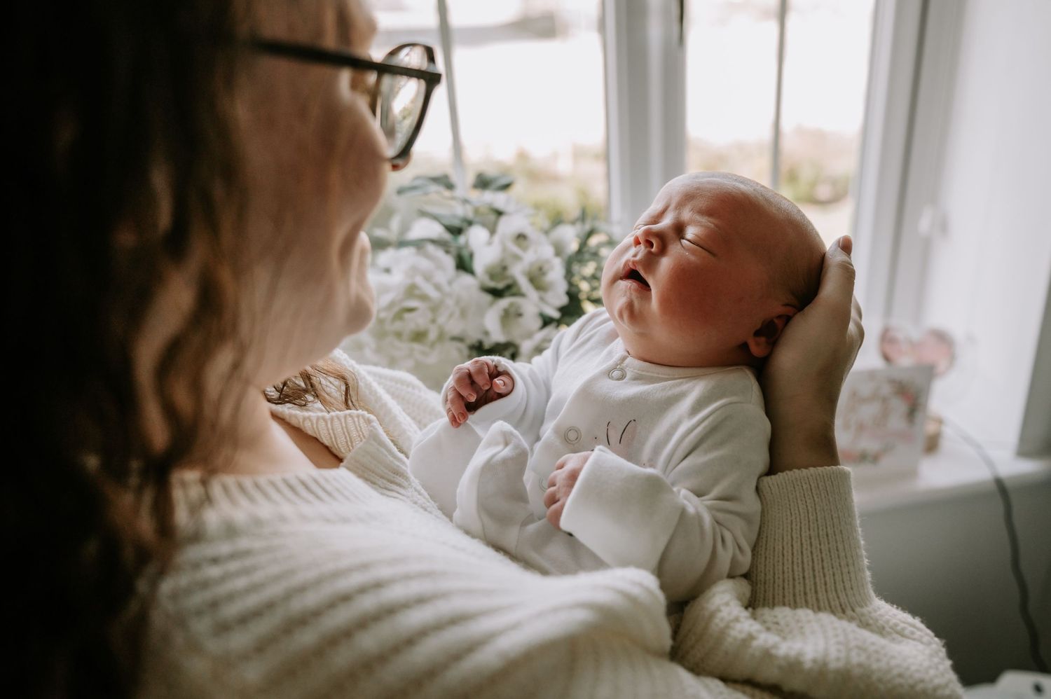 A newborn baby wearing white rests peacefully against a cozy white sweater by a bright window.