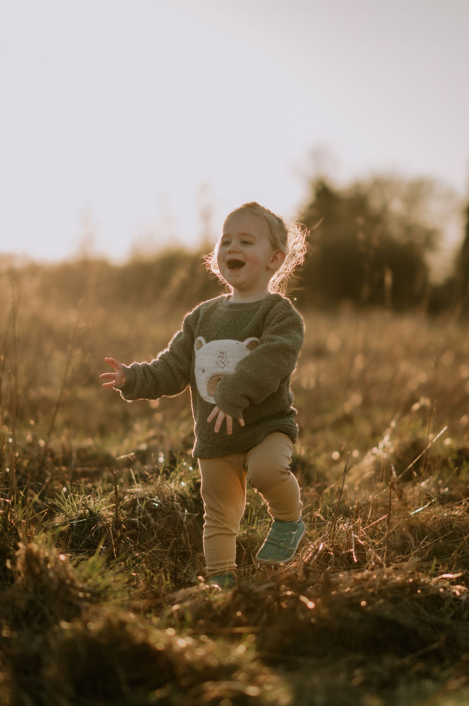 A joyful toddler runs through a golden sunlit meadow at sunset wearing a sweater and knee-high socks.