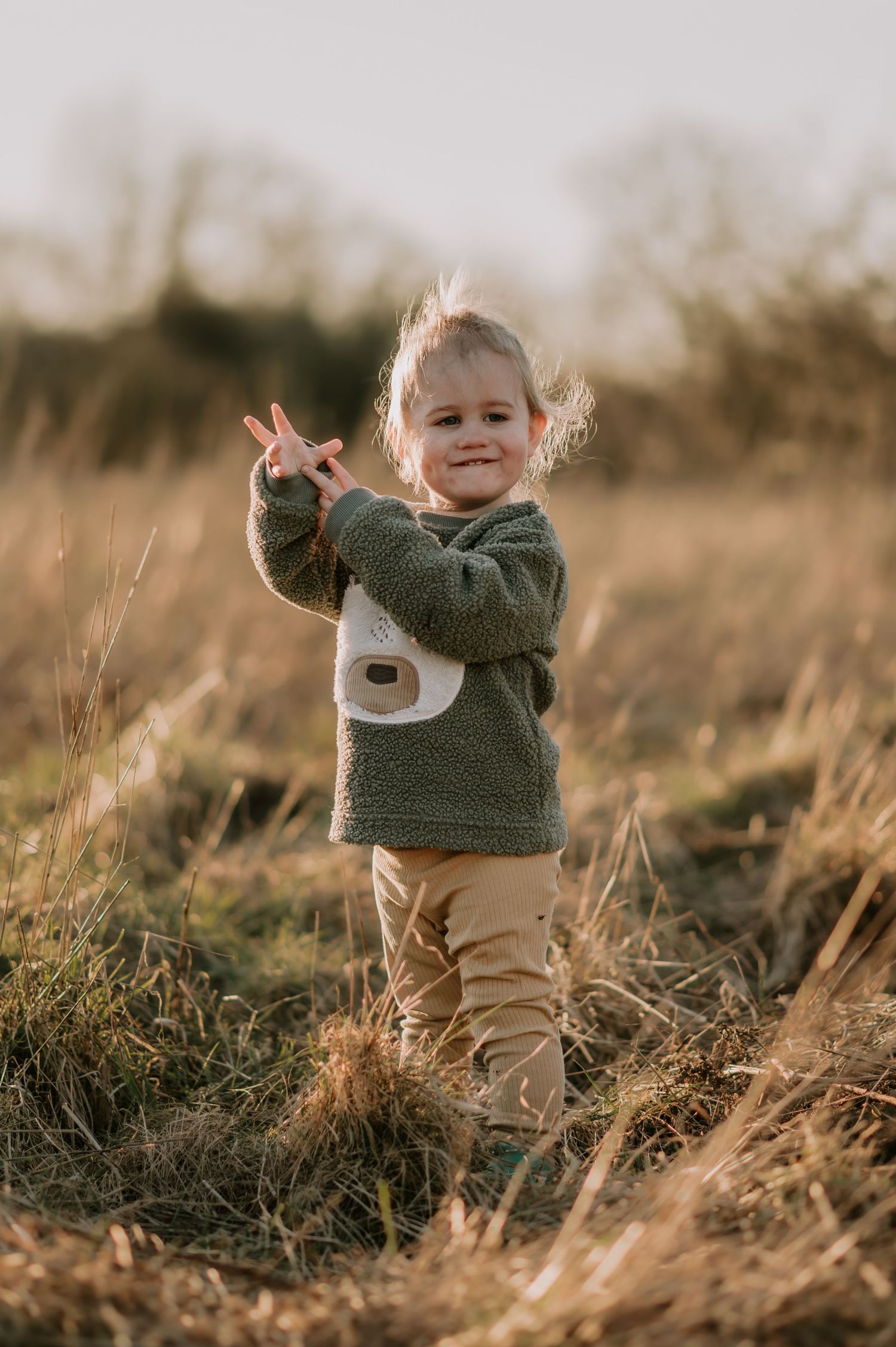 Small child wearing a cozy gray fleece sweater and khaki pants stands in a golden field at sunset.