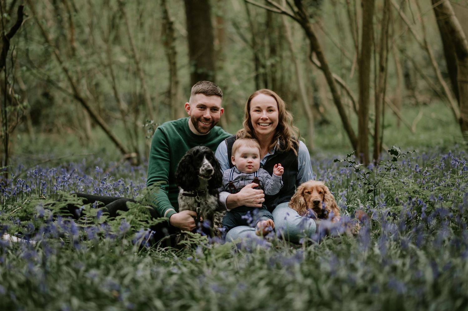 Family and their brown dog sit together in springtime forest surrounded by blooming bluebells.